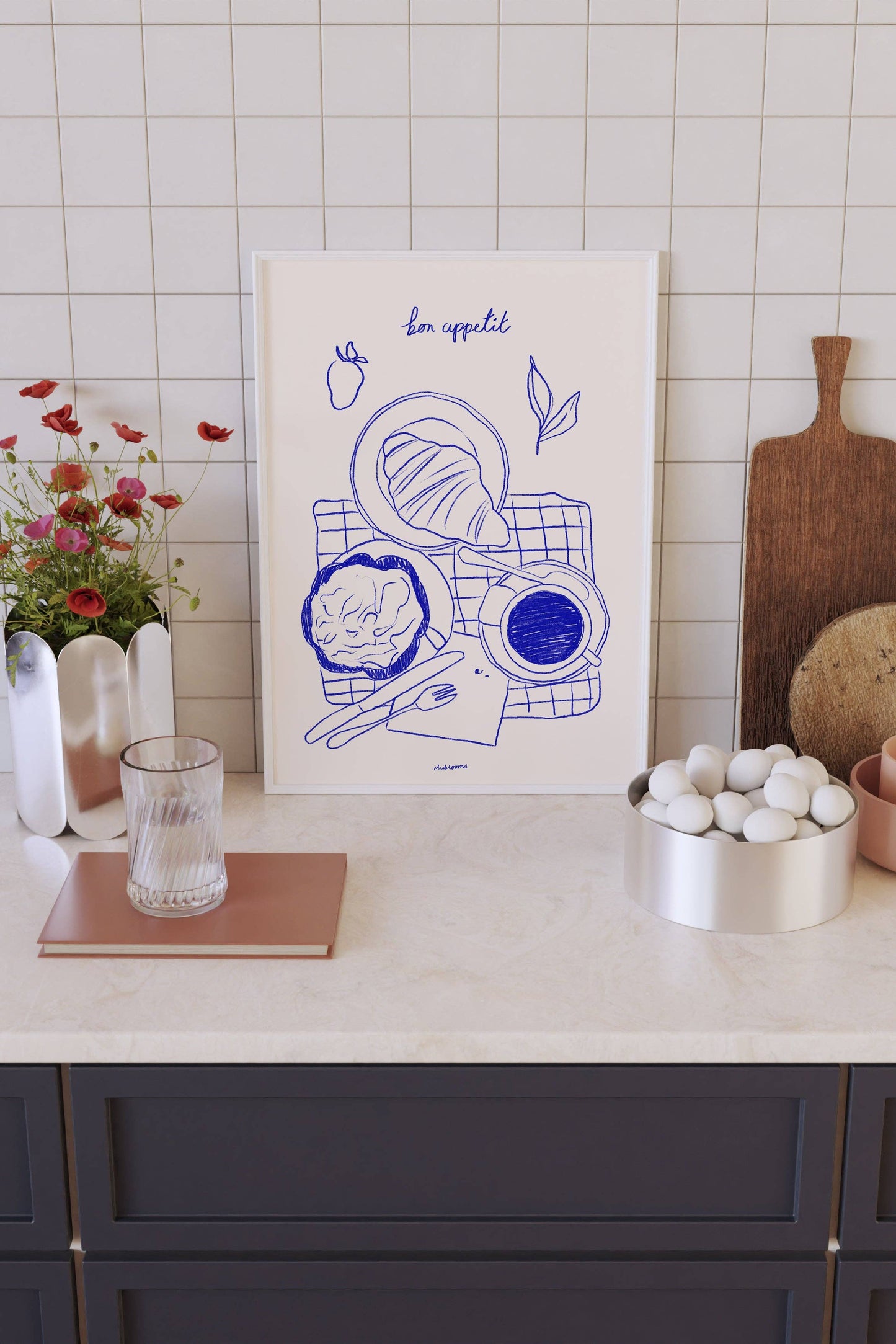 Kitchen counter with a framed artwork, cutting board, and decorative items against a tiled wall.