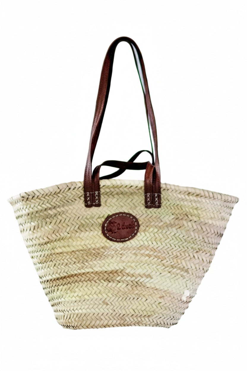 A natural woven straw tote bag with dark brown leather handles and a circular brown leather logo patch on the front, hanging from a wooden peg against a plain white background.