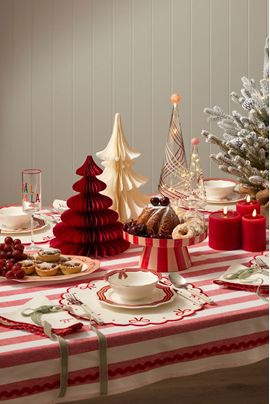 A festive Christmas table setting with a red and white striped tablecloth, red candles, paper Christmas trees, a small flocked tree, and assorted treats on a beige paneled wall background.