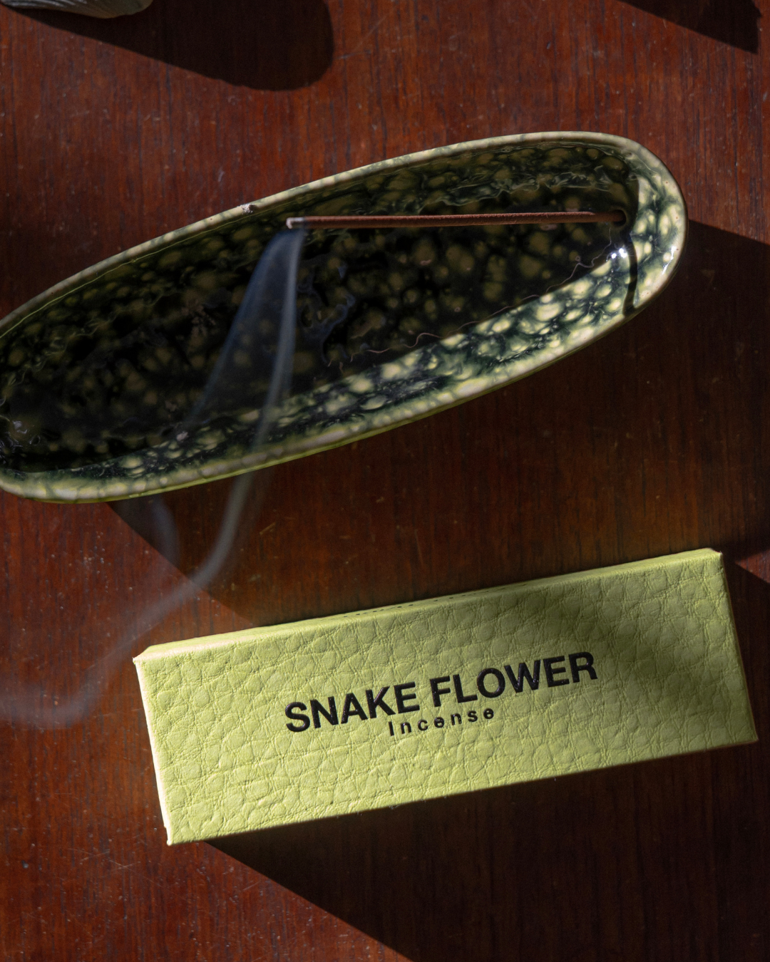 A ceramic incense burner with a black and beige spotted pattern, shown with a pale yellow-green box labeled 'SNAKE FLOWER incense' on a dark wooden surface.