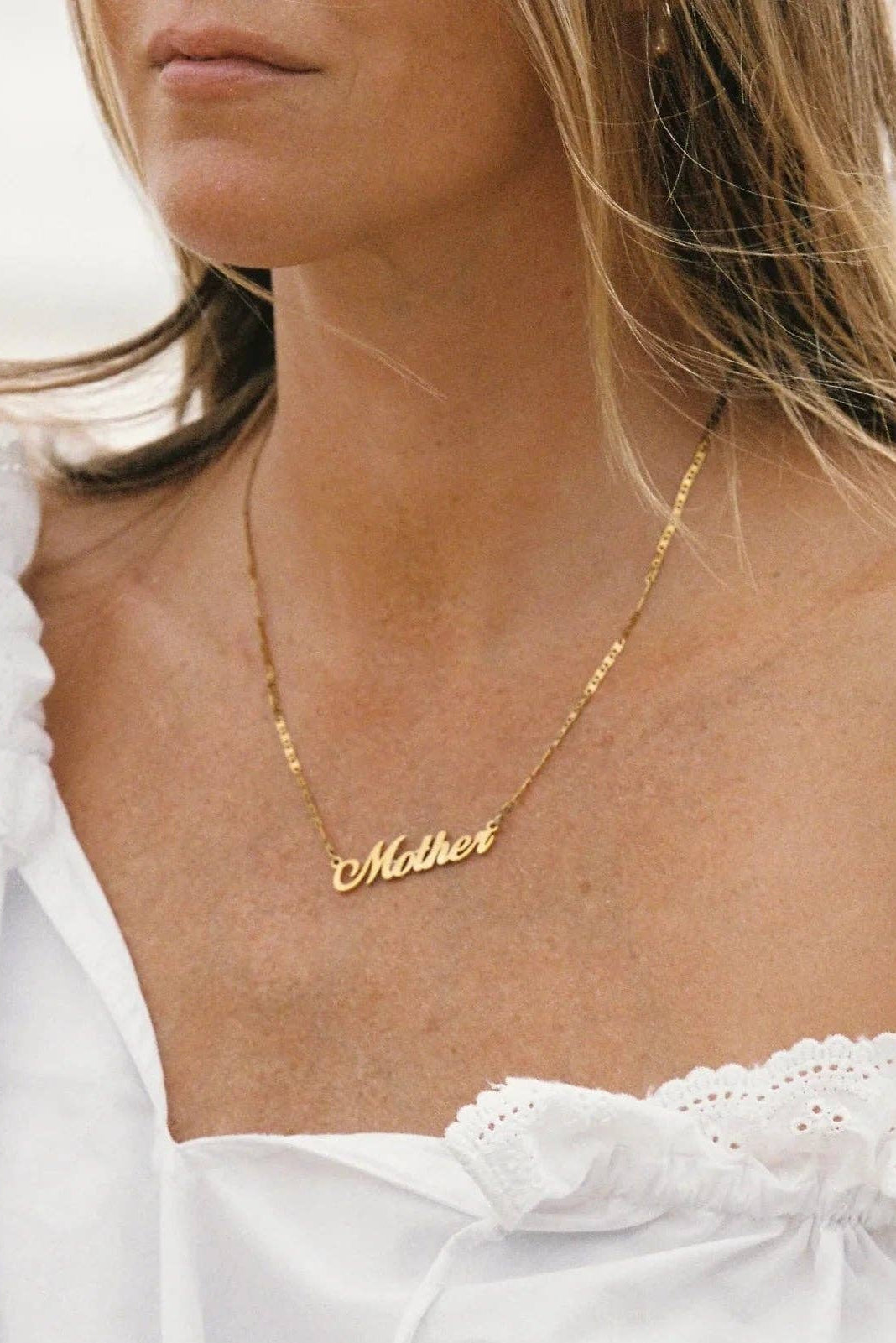 A close-up lifestyle shot of a woman wearing a delicate gold nameplate necklace that reads 'Mother', paired with a white ruffled off-the-shoulder top.