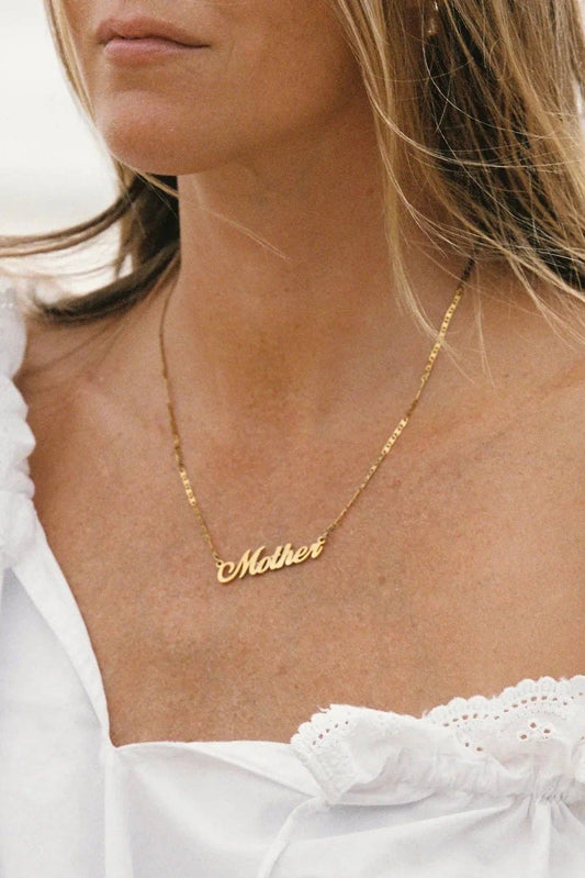 A close-up lifestyle shot of a woman wearing a delicate gold nameplate necklace that reads 'Mother', paired with a white ruffled off-the-shoulder top.