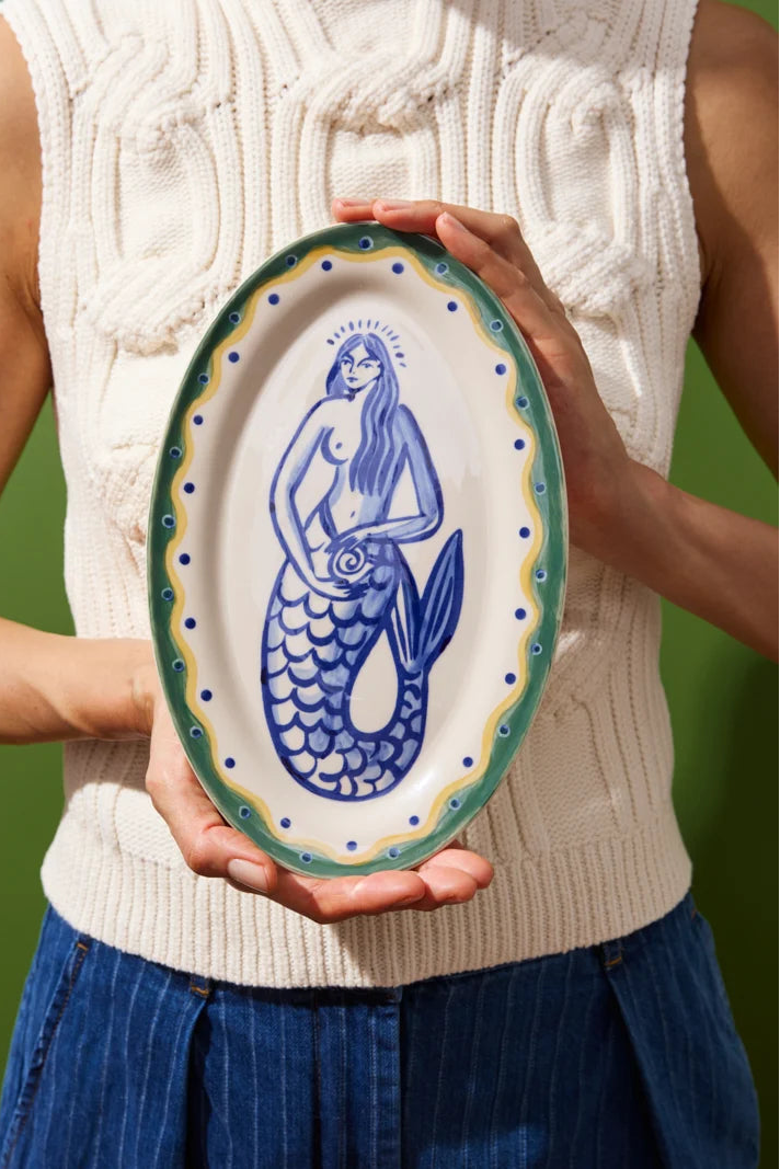 Person holding a ceramic plate with a mermaid design against a green background
