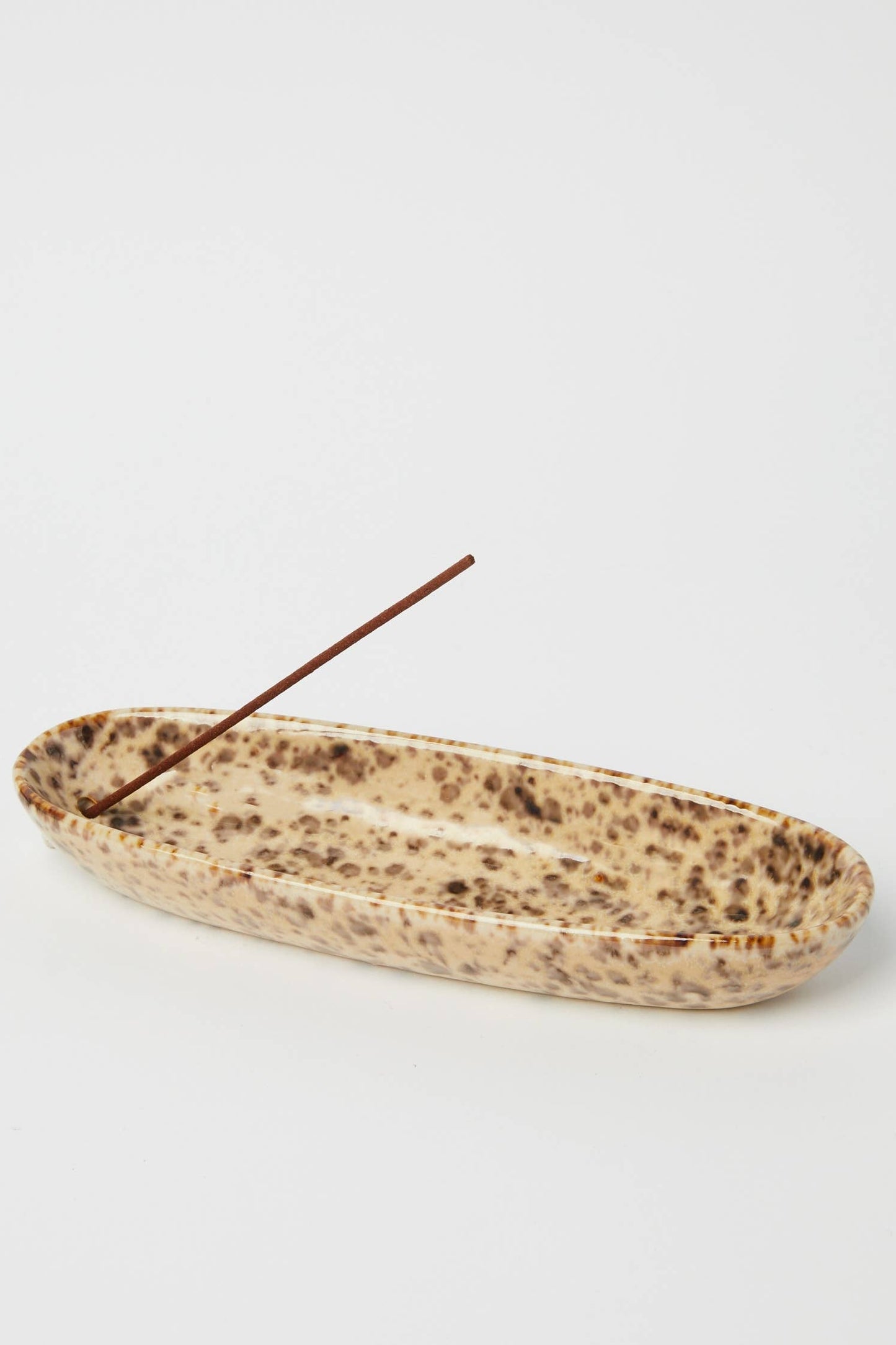A shallow, elongated ceramic dish with a speckled beige and brown glaze, containing a single incense stick, set against a plain light gray background.