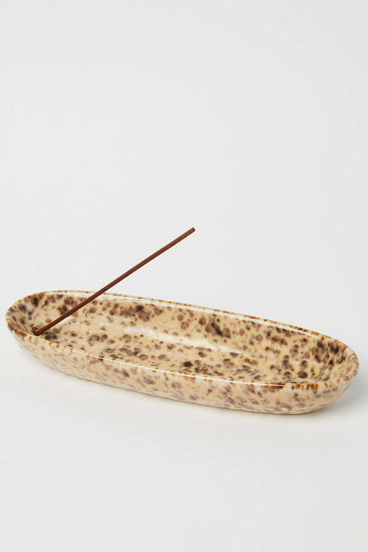 A shallow, elongated ceramic dish with a speckled beige and brown glaze, containing a single incense stick, set against a plain light gray background.