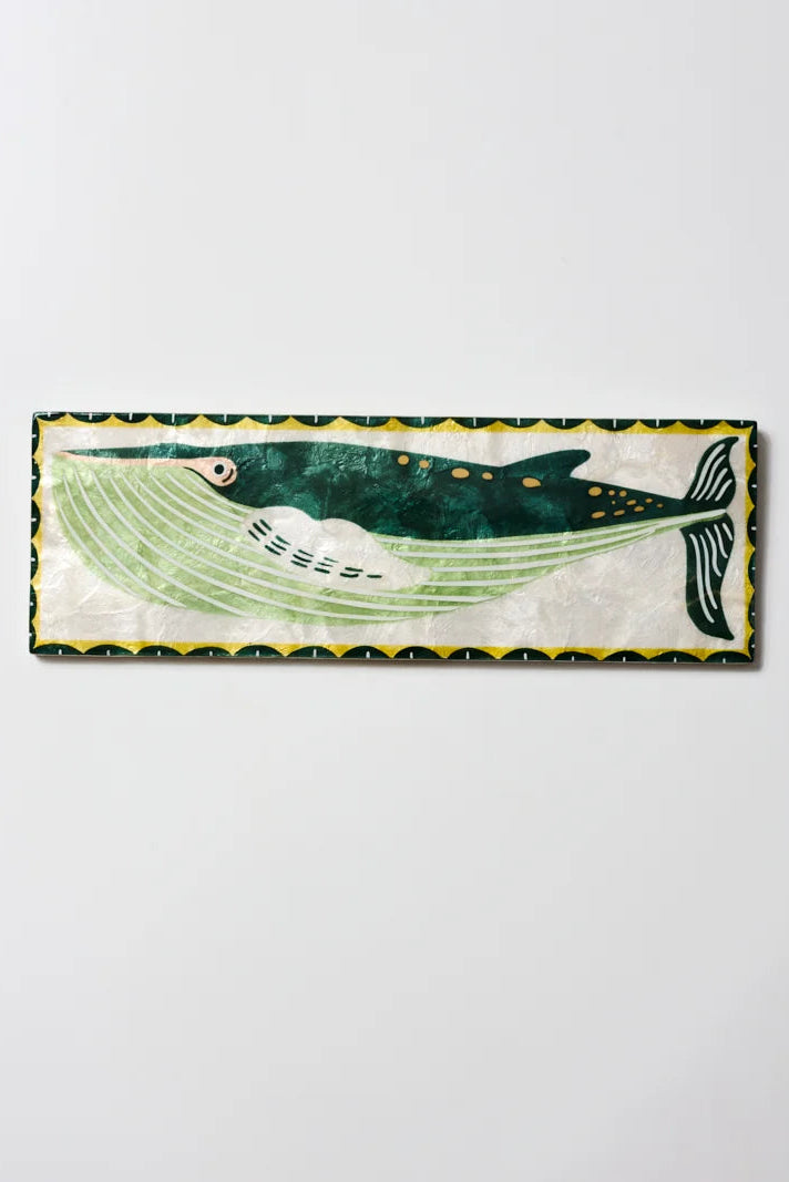 A rectangular decorative wall plaque featuring a stylized whale illustration in green, black, white, and yellow, mounted on a plain light gray wall.