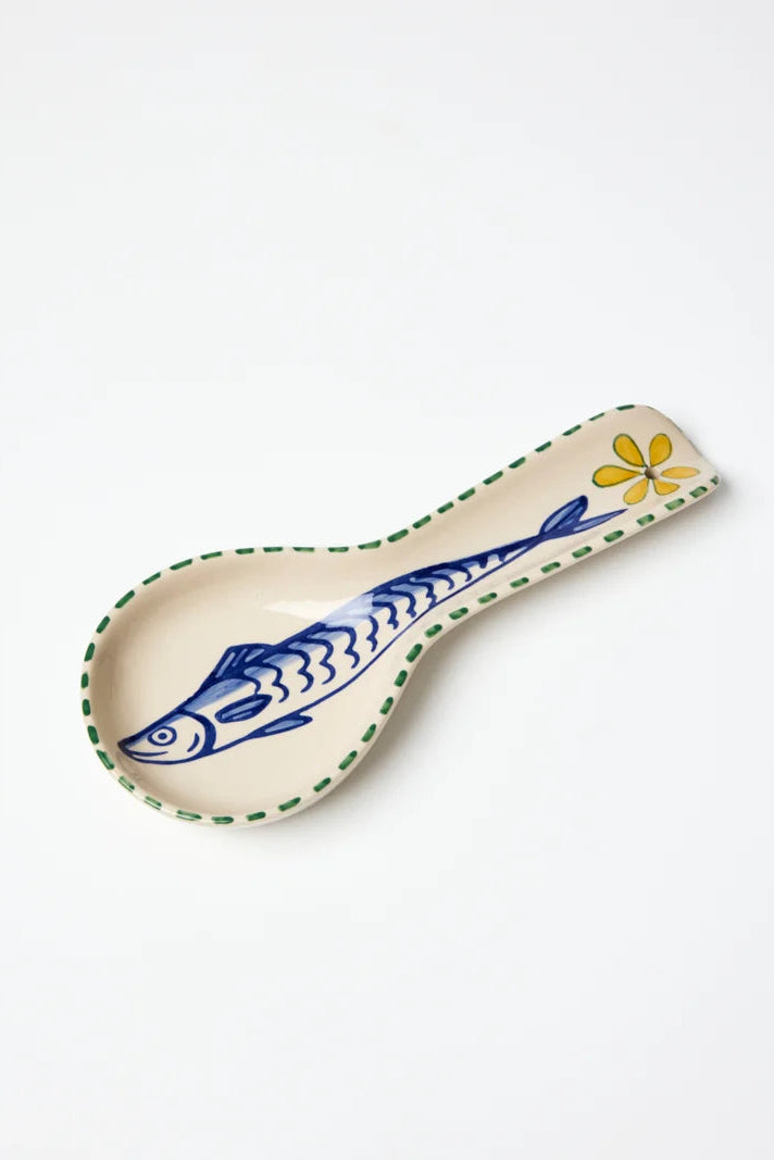 A ceramic spoon rest with a cream-colored base, decorated with a blue sardine fish, a yellow flower, and a green dotted rim, shown on a plain white background.