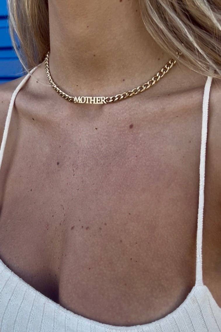 A close-up of a woman wearing a gold chain necklace with the name 'MOTHER' engraved on it, paired with a white ribbed spaghetti-strap top.