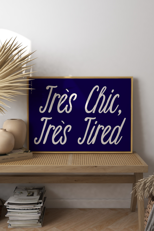 Navy blue sign with white text 'Très Chic, Très Tired' on a wooden shelf with decorative items.