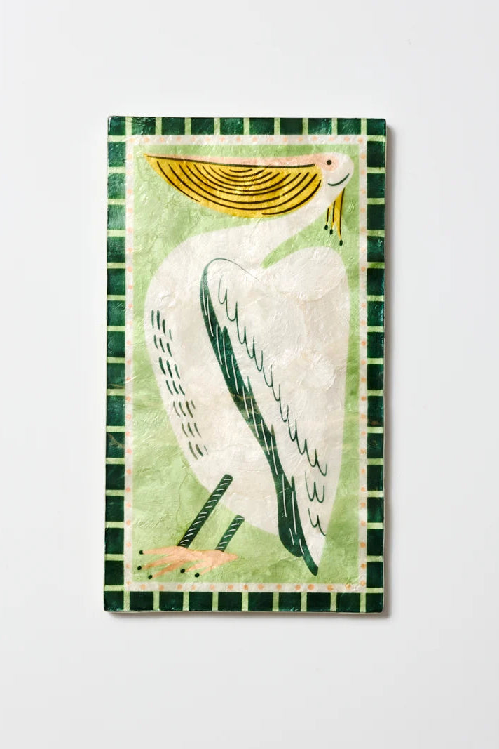 A rectangular decorative tile or plaque featuring a stylized white pelican with a yellow beak on a pale green background, surrounded by a dark green checkered border, shown against a light gray wall.