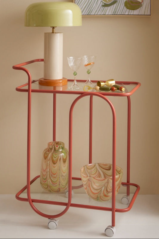 A mid-century modern bar cart in a terracotta red frame with a glass-top shelf and lower shelf, styled with a light green dome lamp, wine glasses, and marbled ceramic vases. The scene is set against a warm beige wall with a framed print, and the text 'COCKTAIL HOUR.