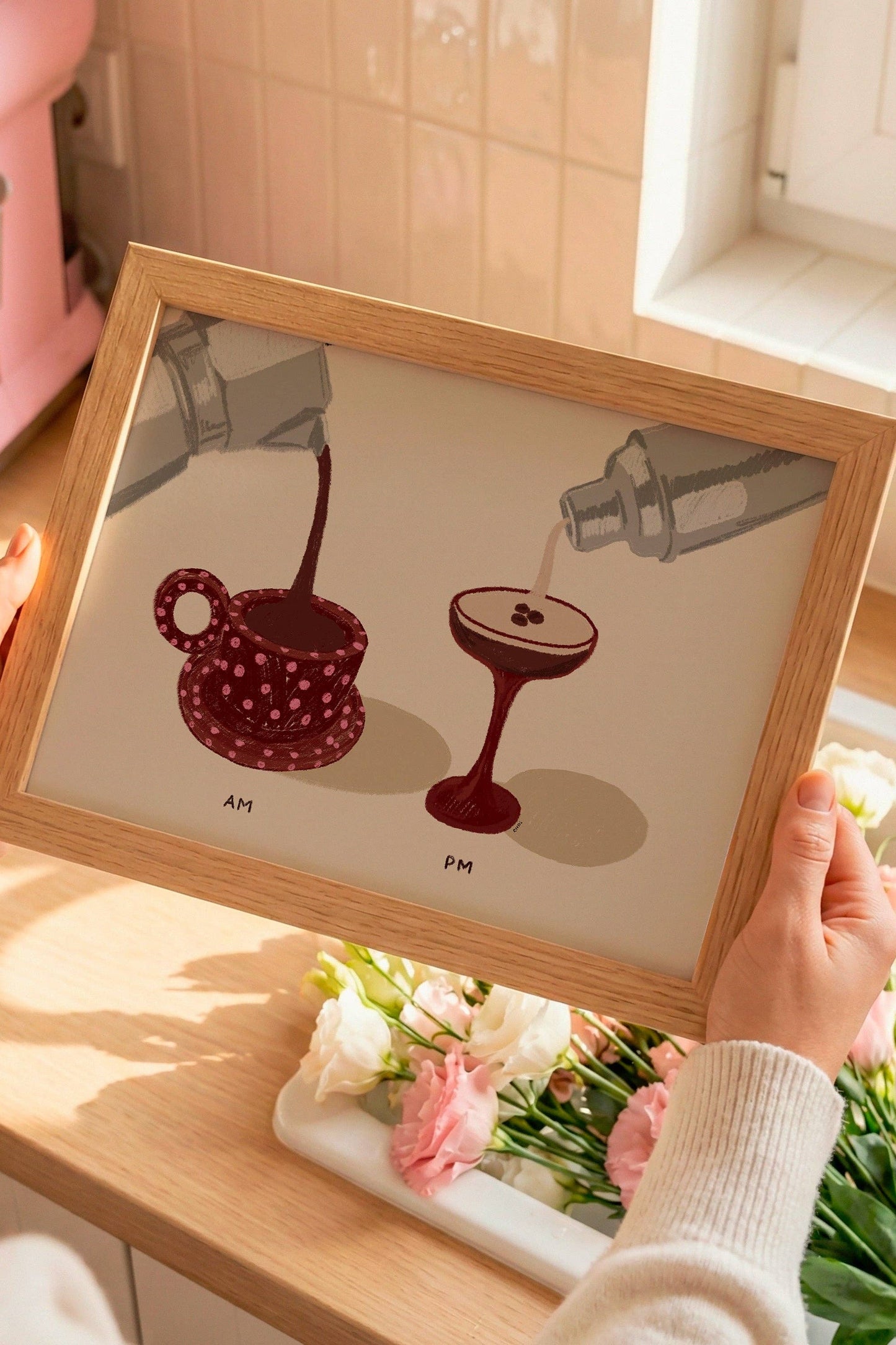 Person holding a framed illustration of a coffee cup and a cocktail on a kitchen counter with a pink mixer in the background.