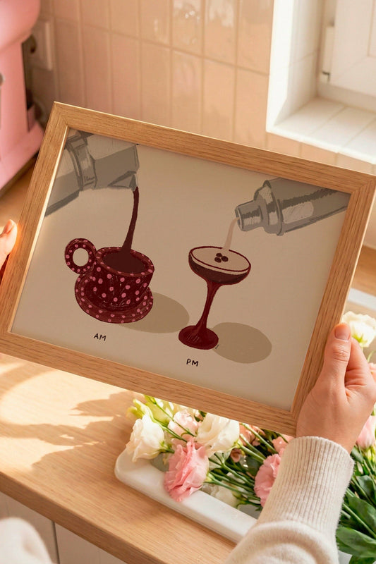 Person holding a framed illustration of a coffee cup and a cocktail on a kitchen counter with a pink mixer in the background.