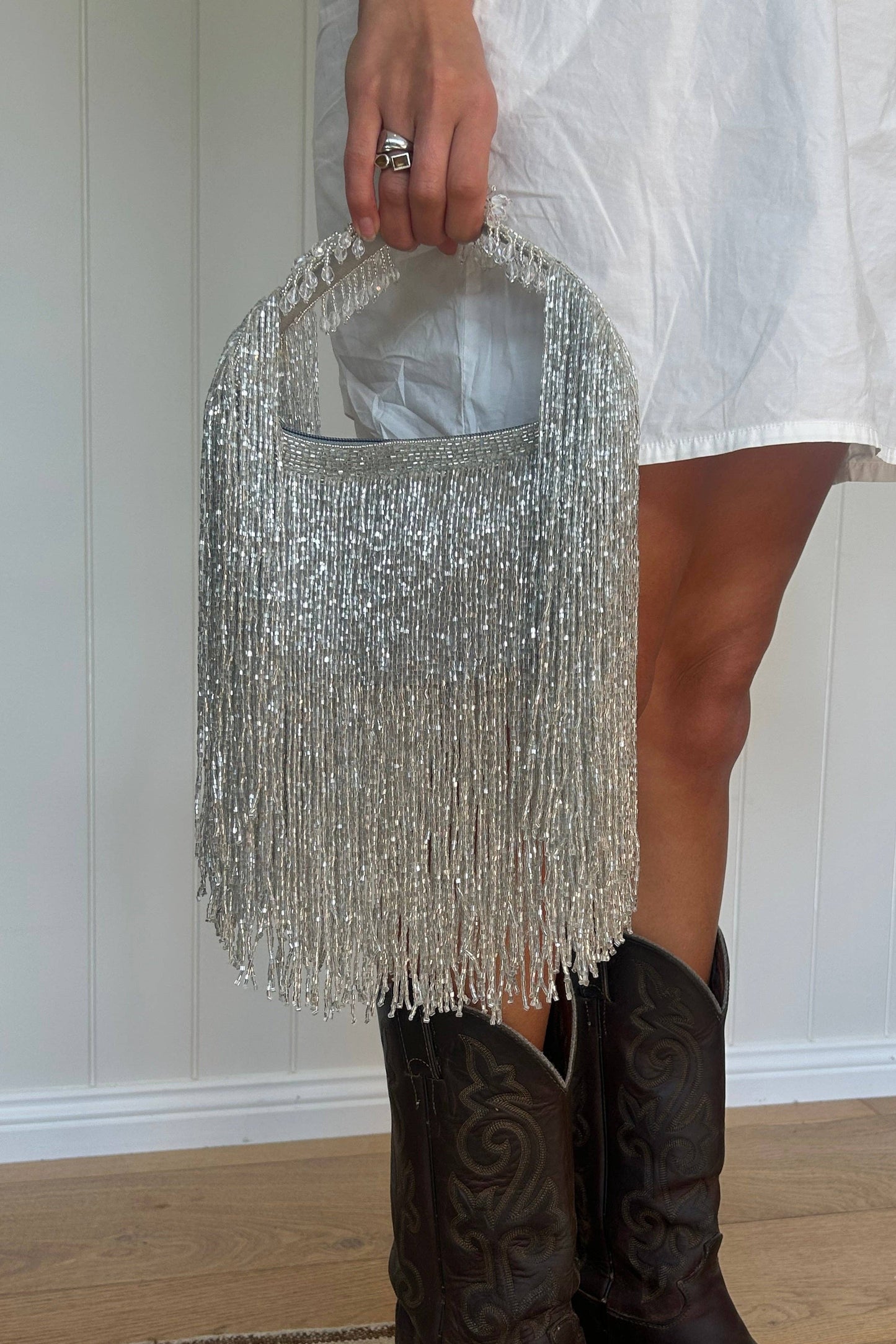 A person wearing a white dress and dark brown western boots holds a large silver beaded fringe handbag in front of a white paneled wall and light wood floor.