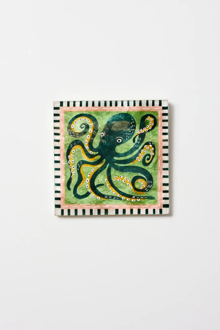 A square decorative art print or tile featuring a whimsical green octopus with yellow tentacles on a light green background, framed by a black-and-white striped border and a pale pink inner edge, shown against a plain white wall.