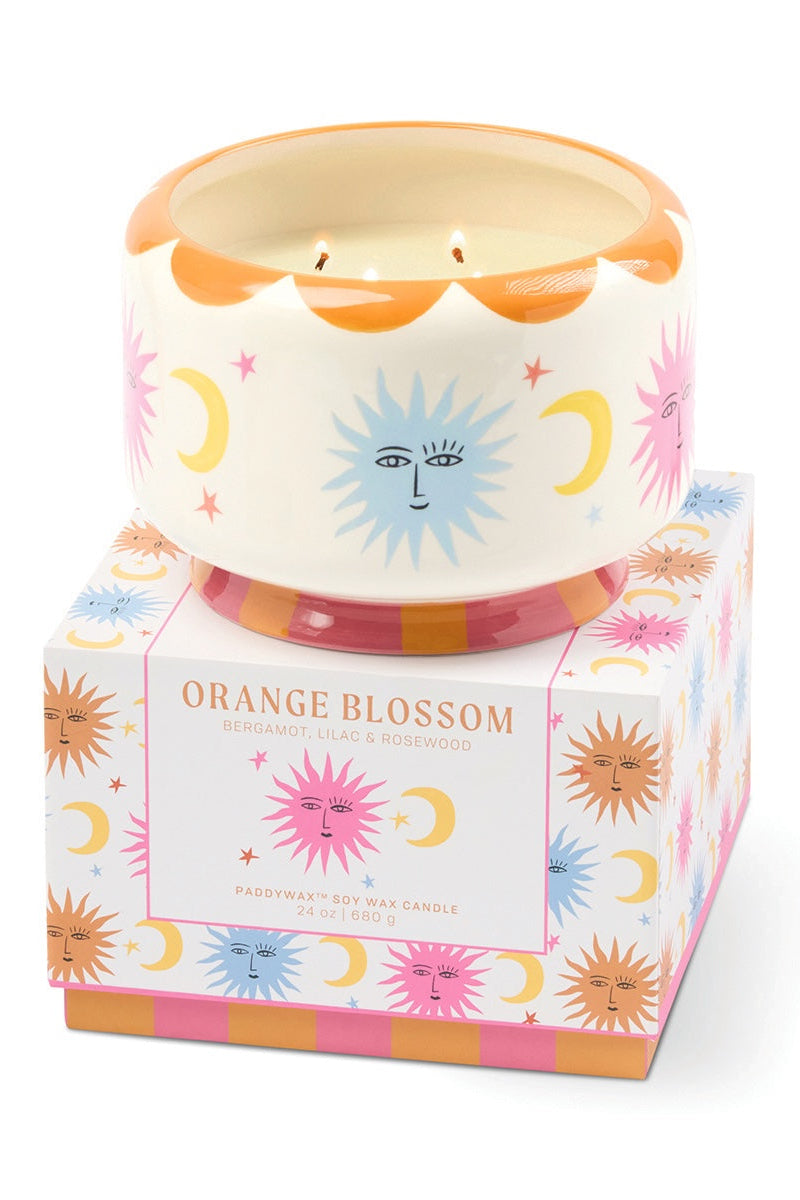 A ceramic candle in a white pot with a scalloped orange rim and a blue sun face design, displayed on top of its illustrated packaging box labeled Orange Blossom by Sweetener.