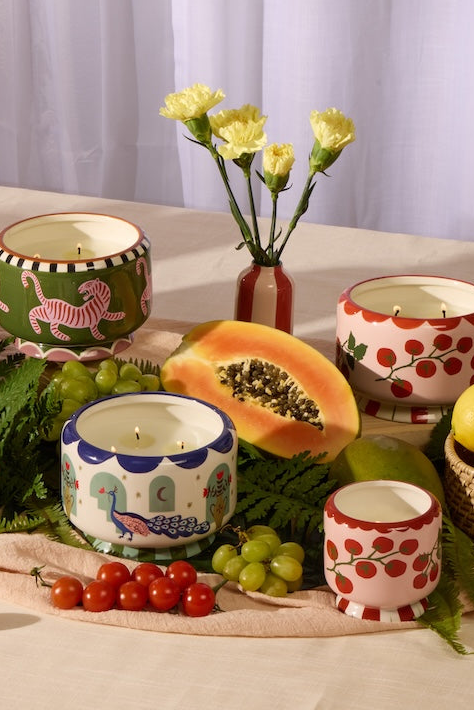 A styled tabletop display of multiple ceramic candle holders with colorful hand-painted patterns, arranged with fresh fruits, limes, and flowers on a cream tablecloth against a white curtain background.