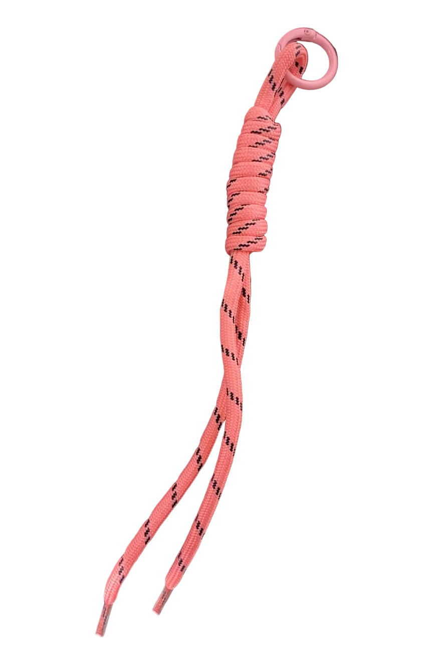 A pink paracord keychain with a black patterned design, a wrapped knot, and a circular ring, resting on a dark brown wooden tray.