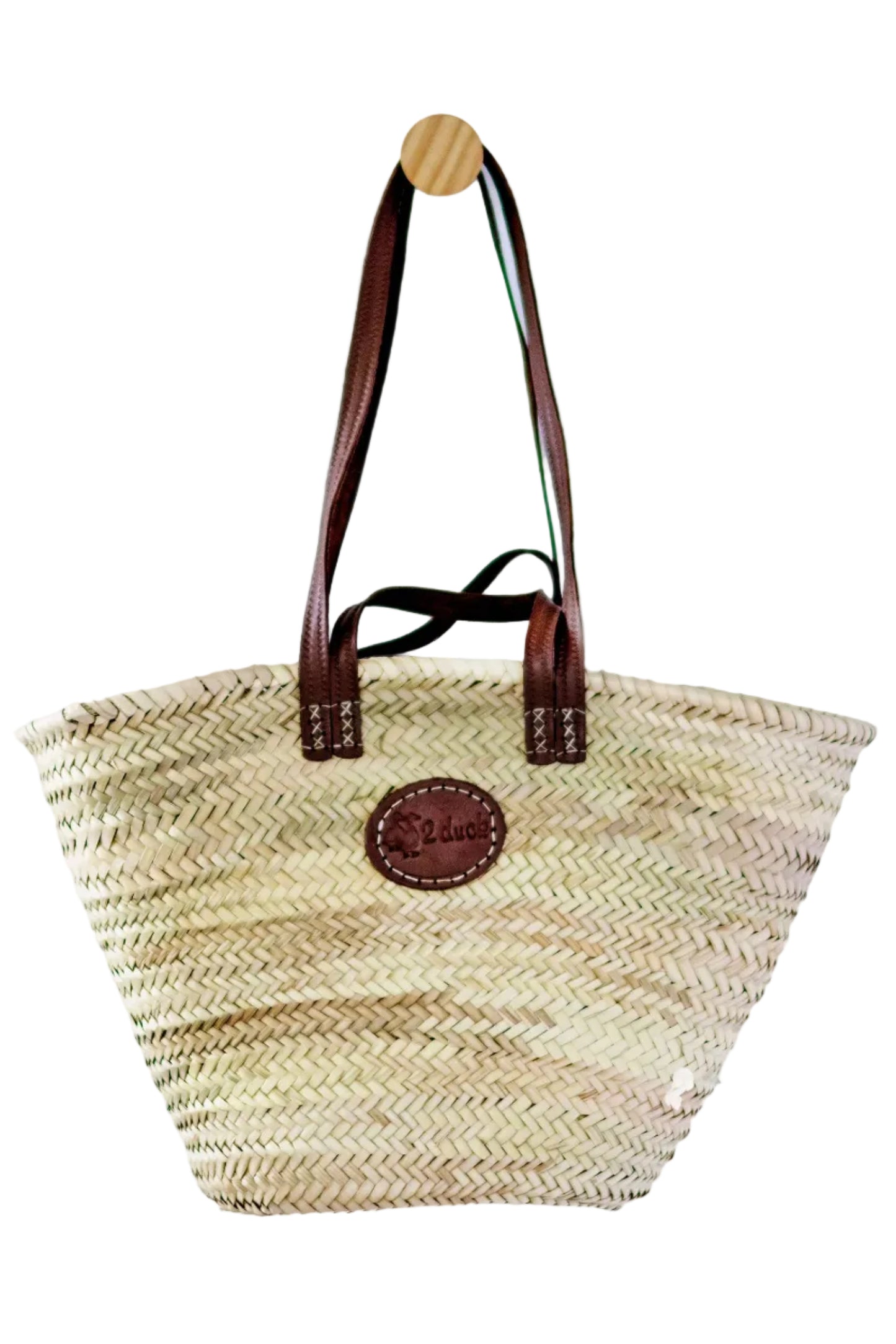 A natural woven straw tote bag with dark brown leather handles and a circular brown leather logo patch on the front, hanging from a wooden peg against a plain white background.
