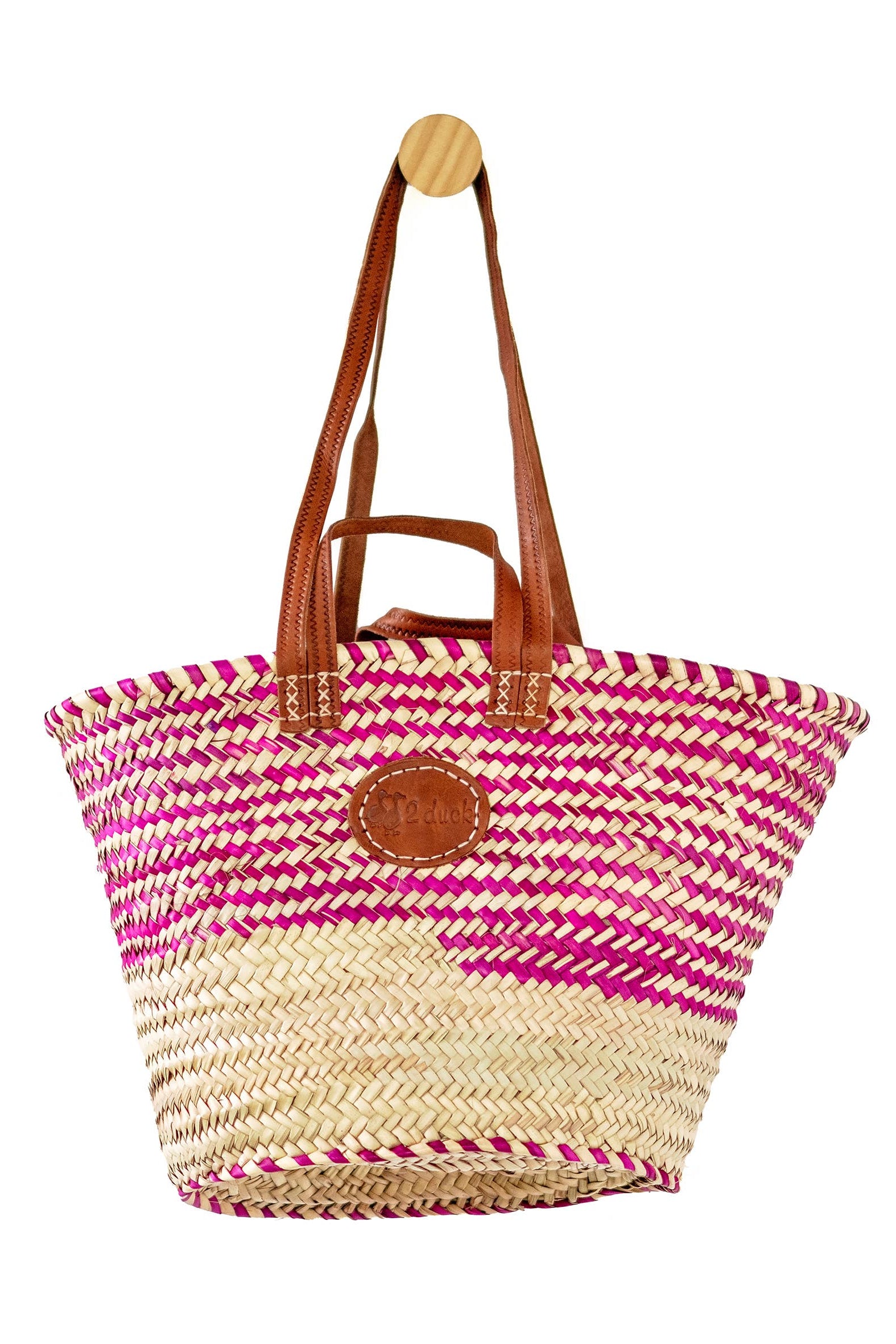 A woven straw tote bag with a pink and beige striped pattern, brown leather shoulder straps, and a round brown leather logo patch on the front, hanging from a wooden peg against a white background.