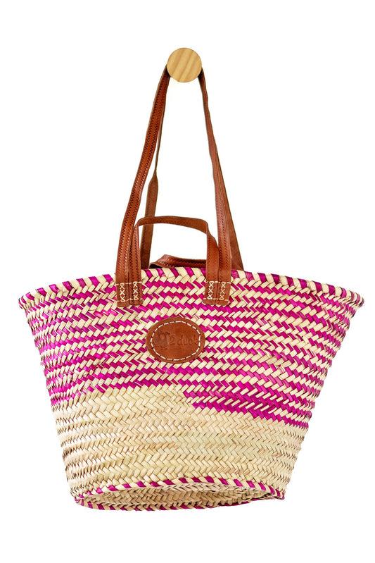 A woven straw tote bag with a pink and beige striped pattern, brown leather shoulder straps, and a round brown leather logo patch on the front, hanging from a wooden peg against a white background.