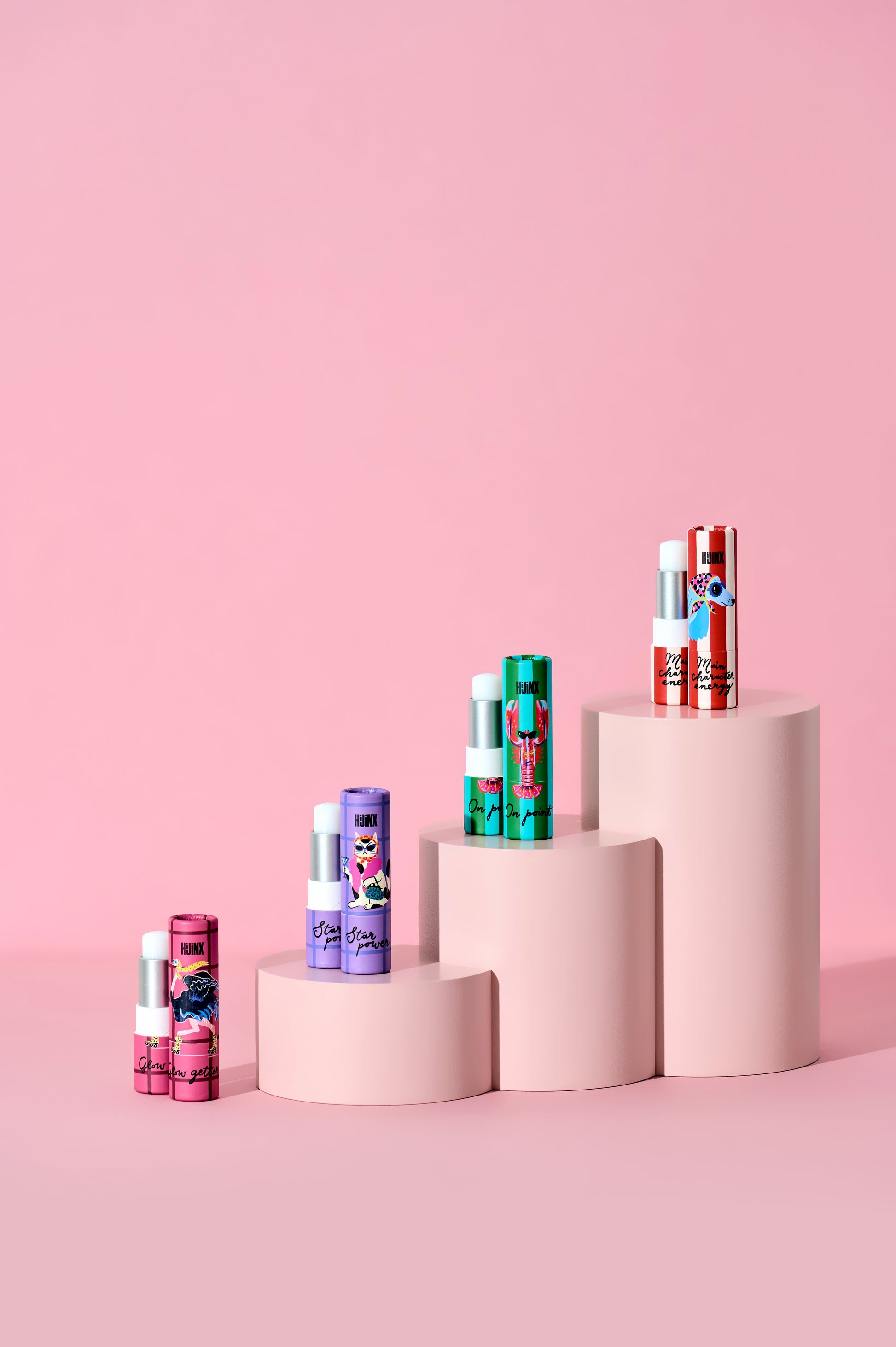 A set of colorful lip balm tubes arranged on a set of pink cylindrical pedestals against a solid pink background. The tubes feature various vibrant colors and cartoon character illustrations.