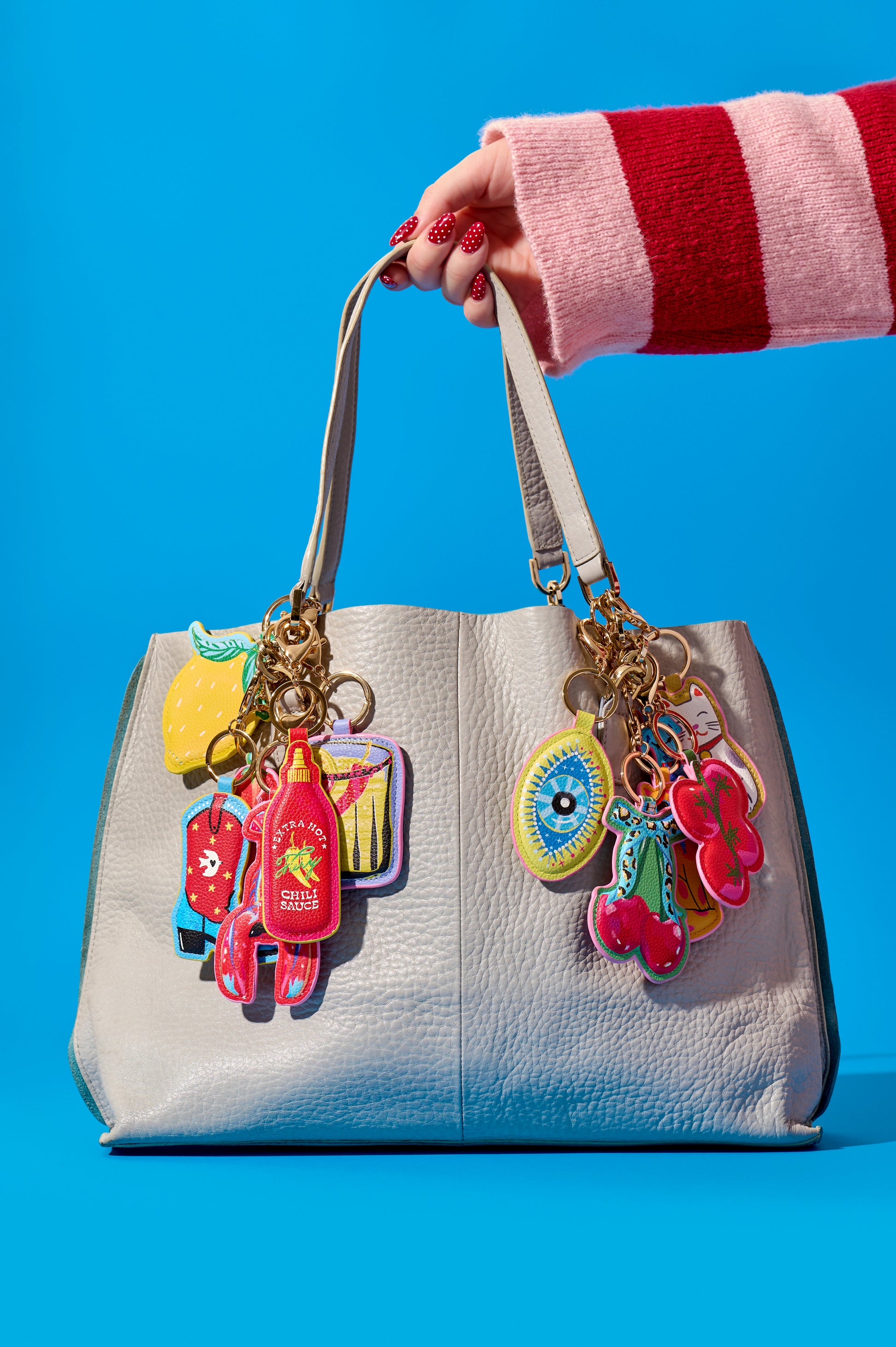 A light gray textured leather handbag with gold-tone hardware and multiple colorful novelty charms, held by a hand wearing a pink and red striped sweater against a solid blue background.