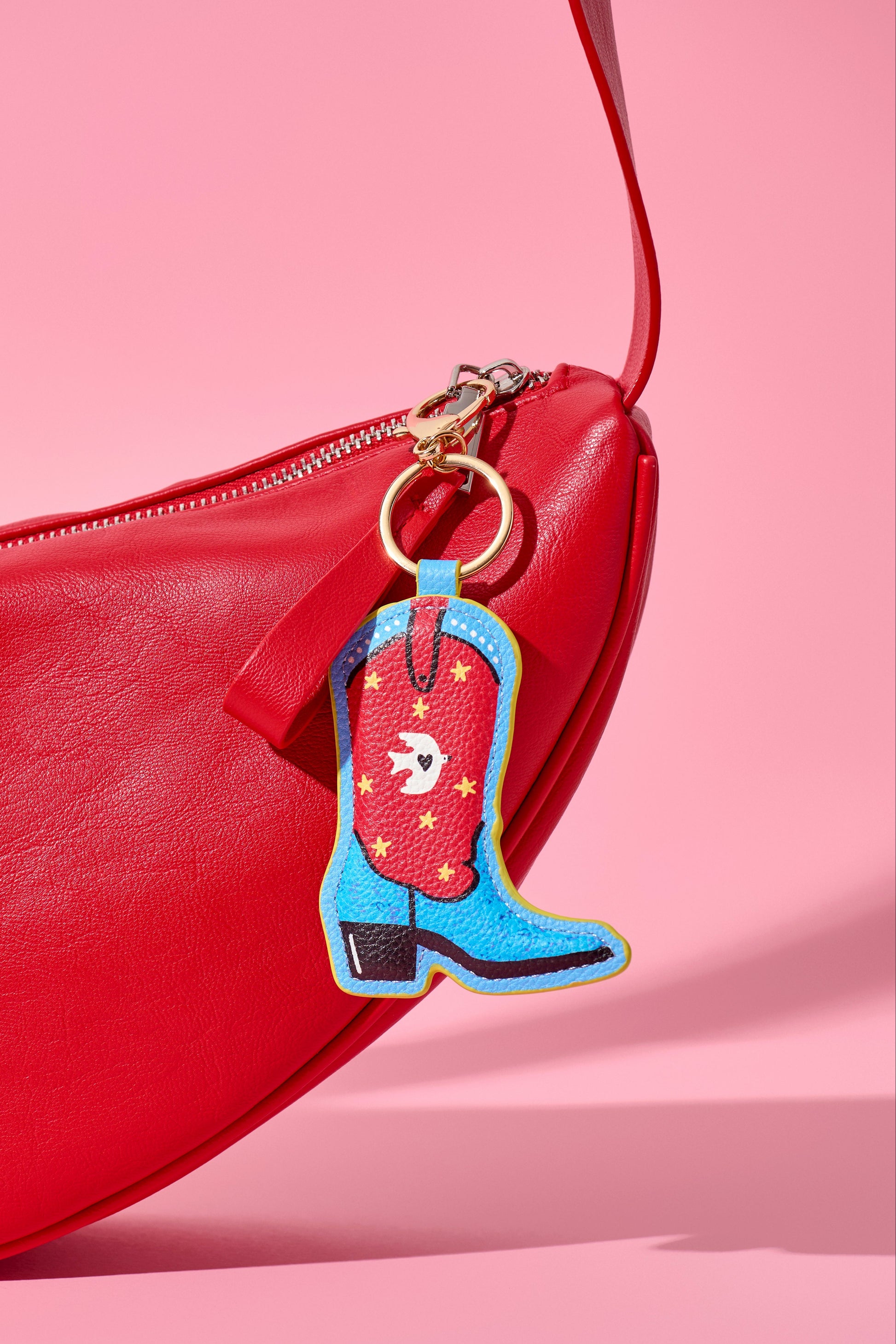 A close-up of a bright red leather bag with a zipper, featuring a colorful embroidered cowboy boot charm in blue, red, yellow, and black attached by a gold ring, set against a solid pink background.