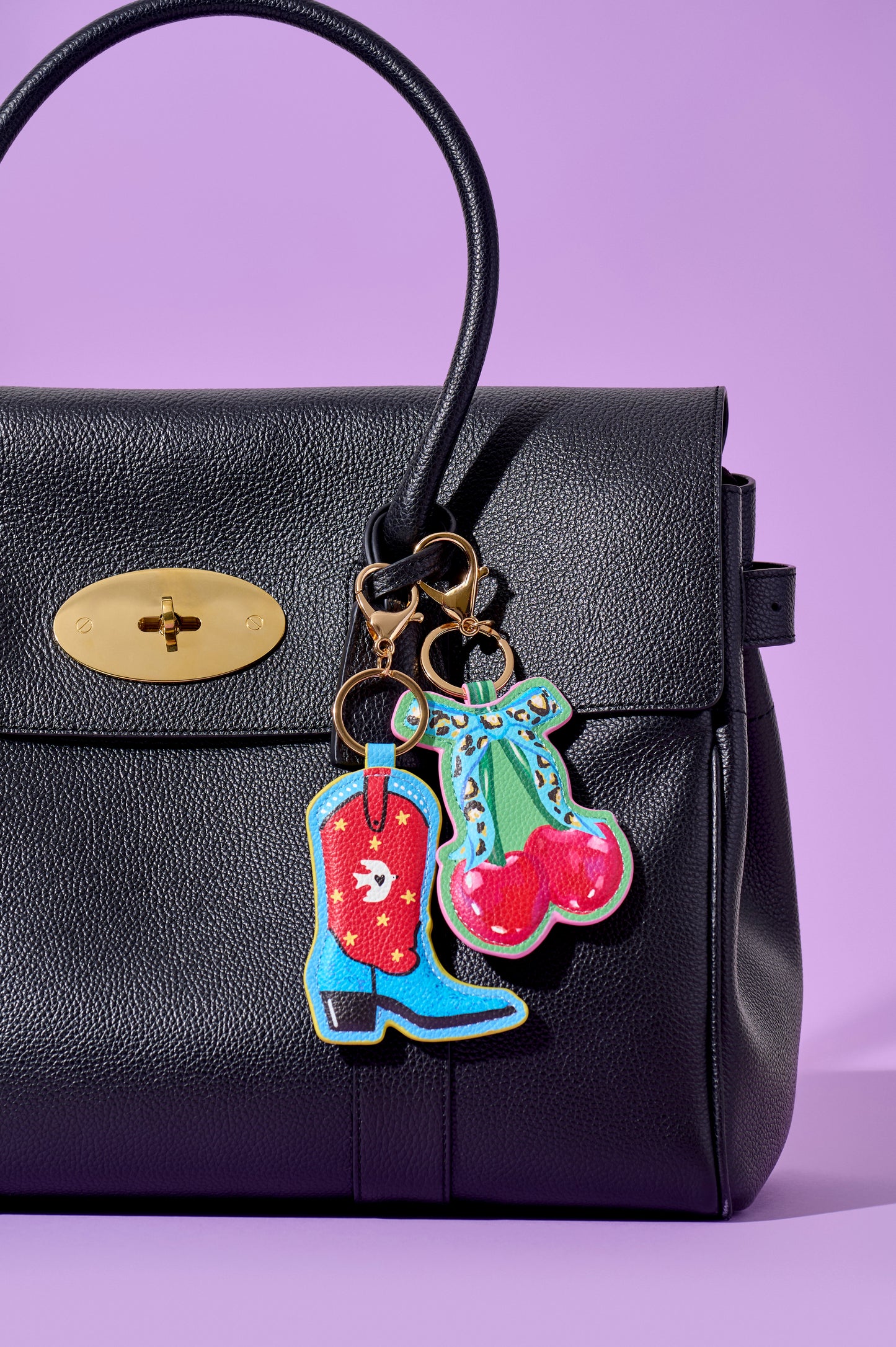 A black textured leather handbag with a gold turn-lock clasp, featuring colorful embroidered cowboy boot and cherry keychain charms hanging from gold-tone rings, set against a solid lavender background.