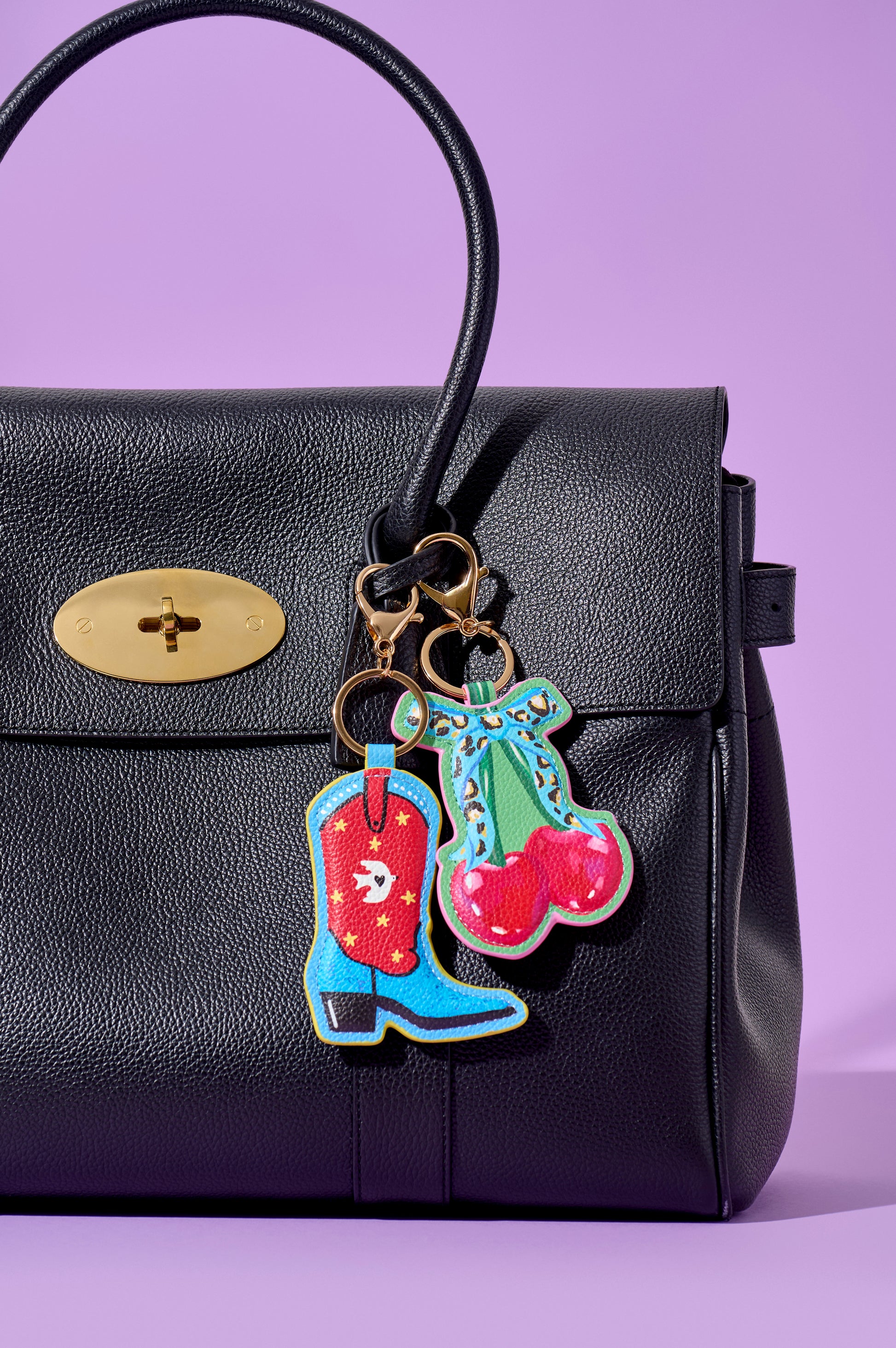 A black textured leather handbag with a gold turn-lock clasp, featuring colorful embroidered cowboy boot and cherry keychain charms hanging from gold-tone rings, set against a solid lavender background.