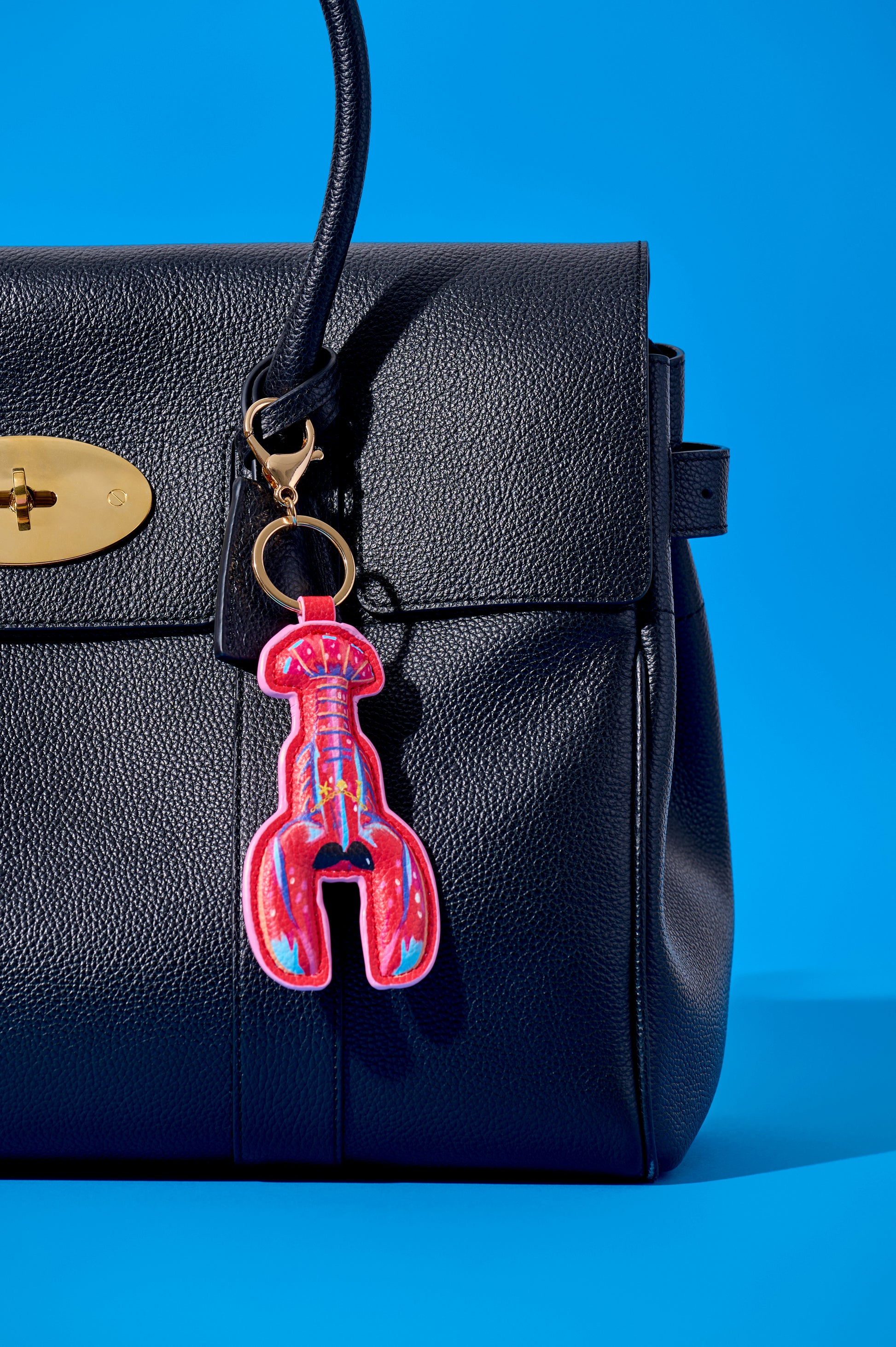 A close-up of a black pebbled leather handbag with a gold turn-lock closure and a lobster-shaped keychain charm in bright pink, red, and blue, set against a solid blue background.