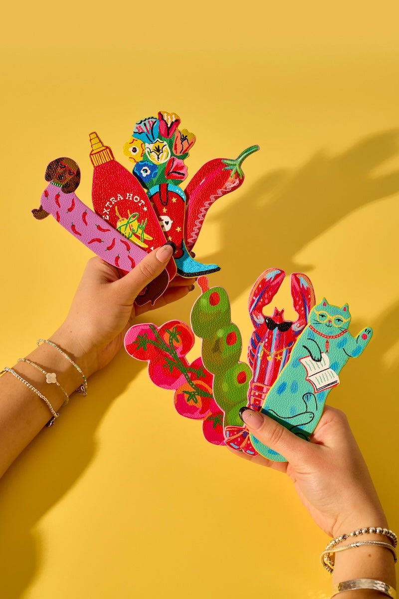 Two hands holding colorful, handcrafted paper cutout bookmarks against a solid yellow background. The bookmarks feature whimsical illustrations including a pink strawberry donut, red hot dogs, a green chili pepper, a red pepper with a blue cat reading a book, and a red pepper with a pink cat.