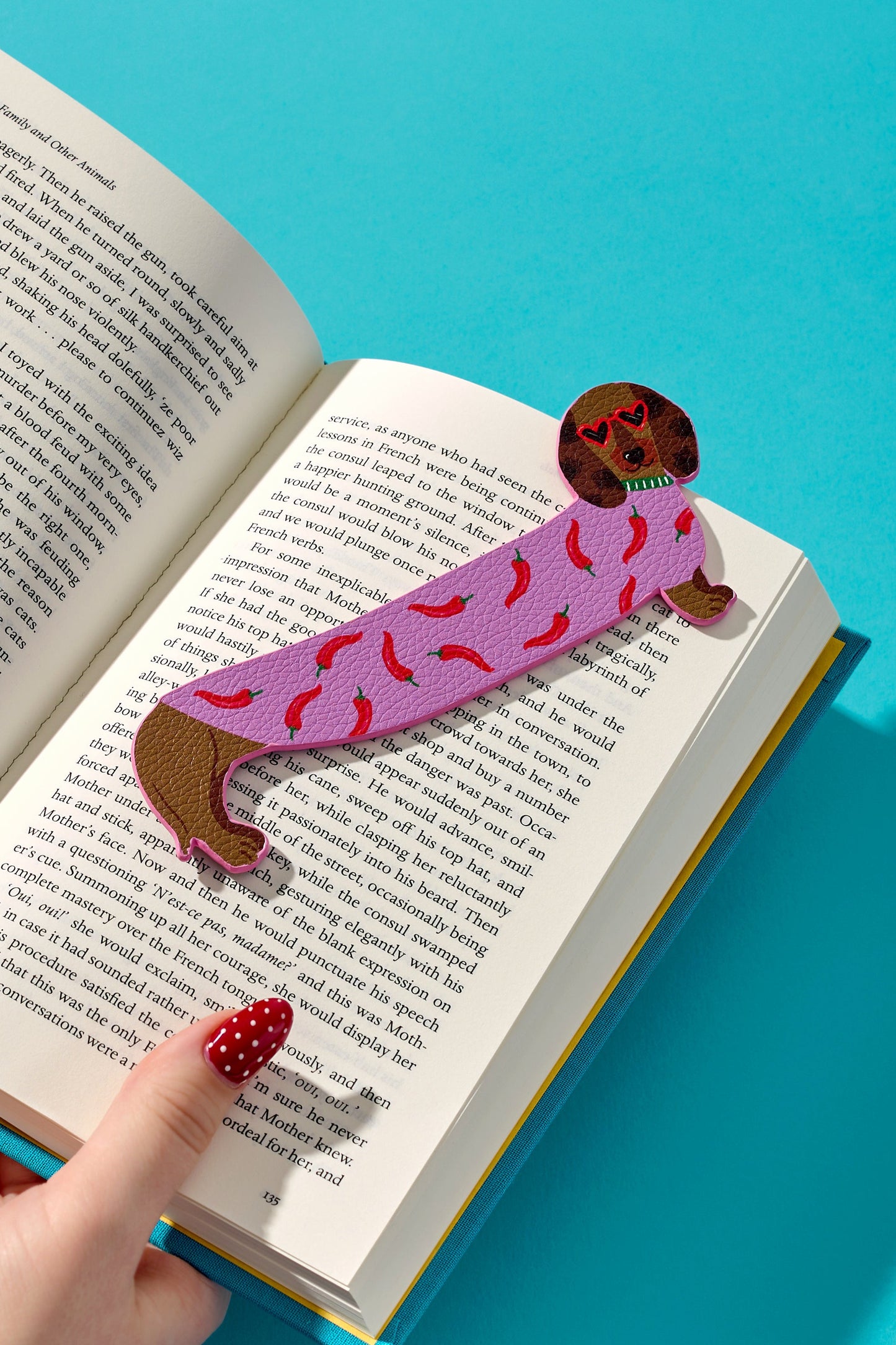 A pink and brown dog-shaped bookmark with sprinkles rests on the pages of an open book held by two hands against a bright blue background.