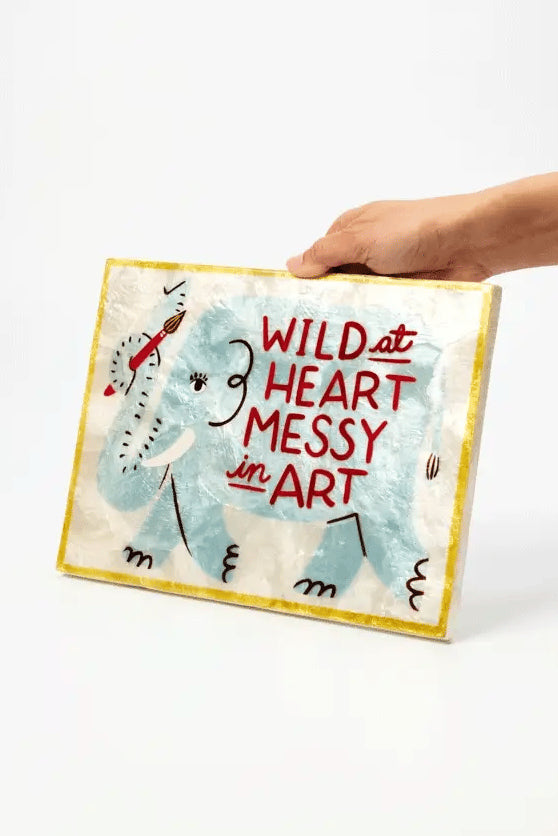 A hand holds a square canvas art print featuring a childlike blue elephant and the text 'WILD at HEART MESSY in ART' against a plain white background.