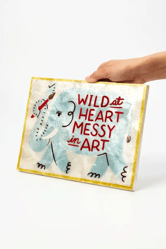 A hand holds a square canvas art print featuring a childlike blue elephant and the text 'WILD at HEART MESSY in ART' against a plain white background.