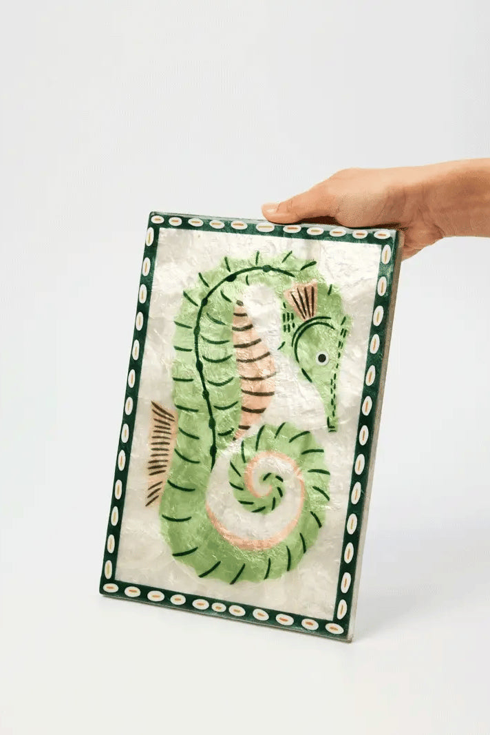 A hand holding a rectangular decorative tile featuring a stylized green seahorse on a white background with a dark green patterned border.
