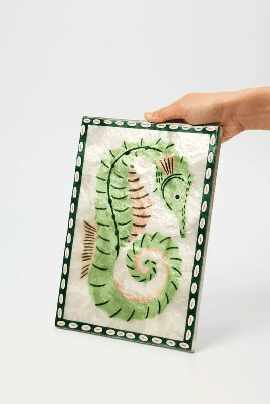 A hand holding a rectangular decorative tile featuring a stylized green seahorse on a white background with a dark green patterned border.
