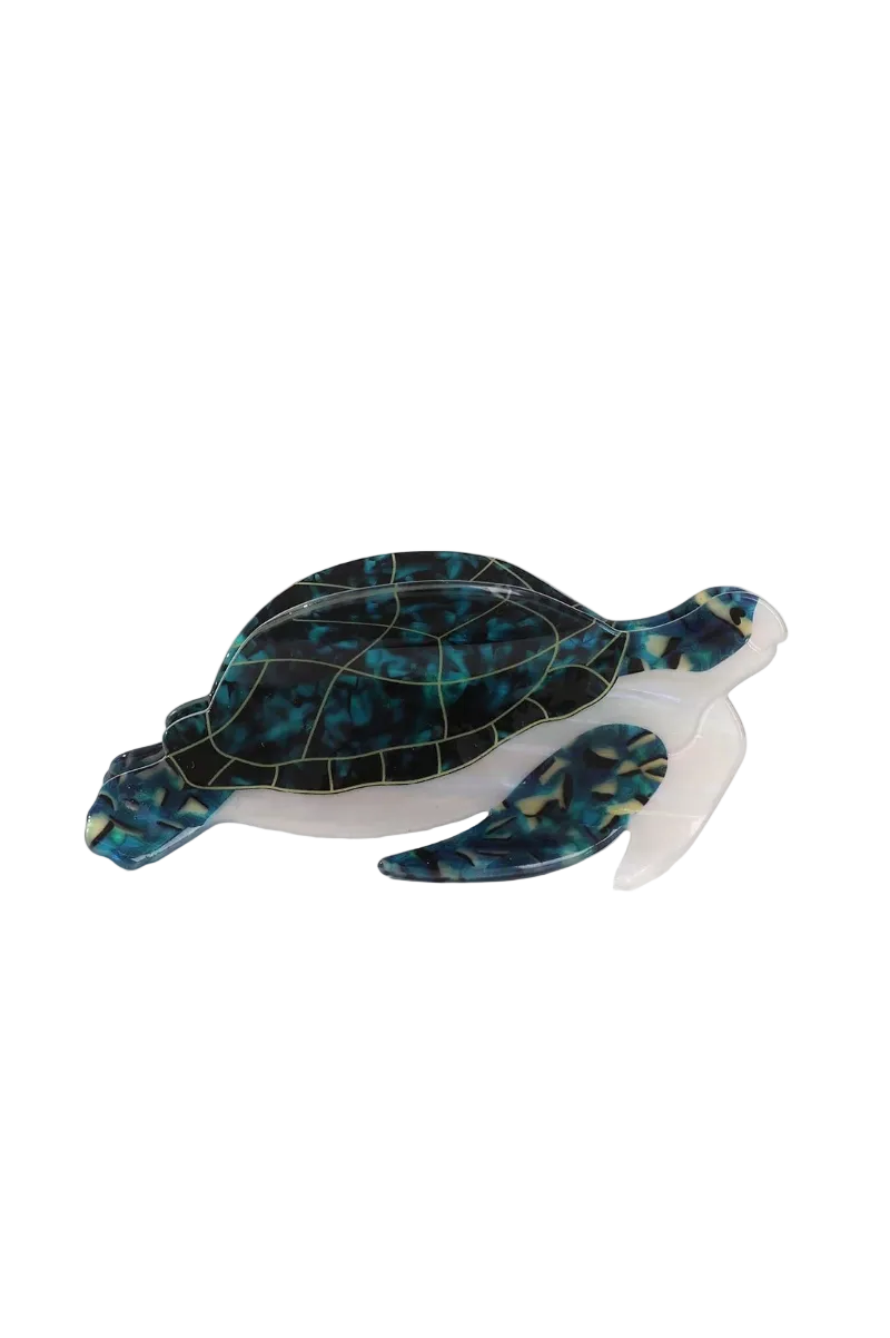 A translucent turtle-shaped hair claw clip with a marbled black and teal resin shell and a clear translucent body, displayed on a white surface with a neutral woven prop in the background.