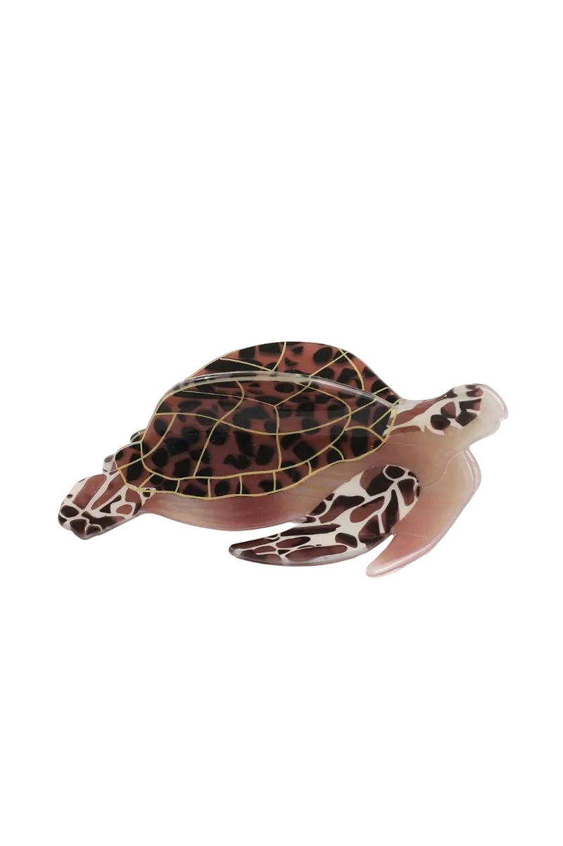 A translucent resin sea turtle hair claw with a brown and black patterned shell and pinkish-brown body, displayed on a clean white surface with a soft beige fabric backdrop.