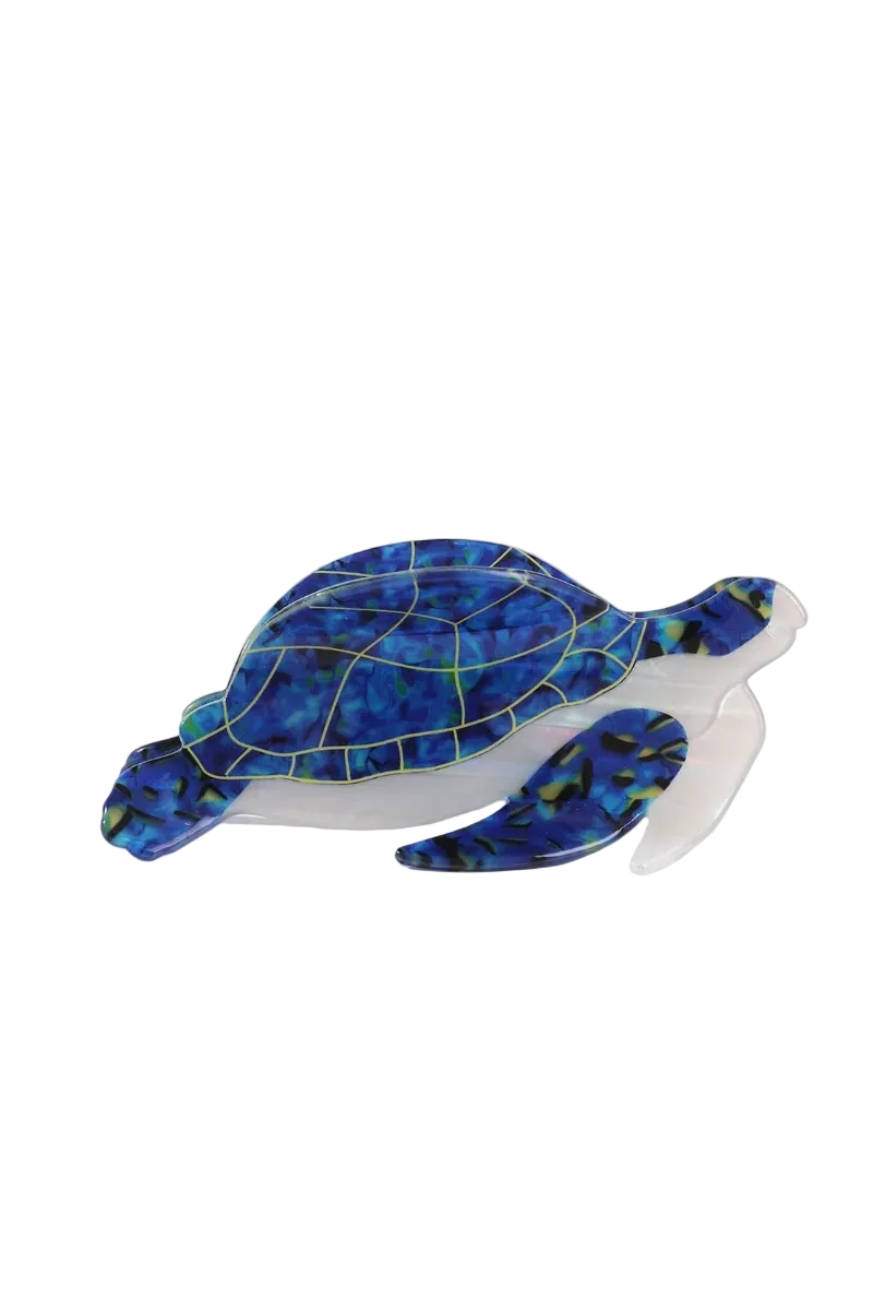 A translucent sea turtle hair claw with a blue and black marbled shell and a clear body, styled on a white surface with a beige woven mat in the background.