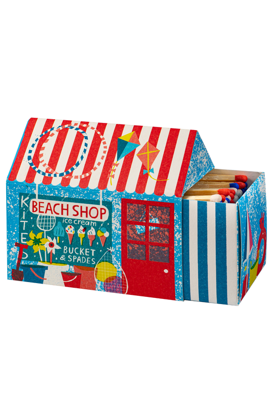 A decorative matchbox shaped like a beach shop tent, featuring red and white stripes on the roof, a blue side panel with 'BEACH SHOP' and ice cream graphics, and a striped front panel. The open top reveals wooden matches with red and blue tips.
