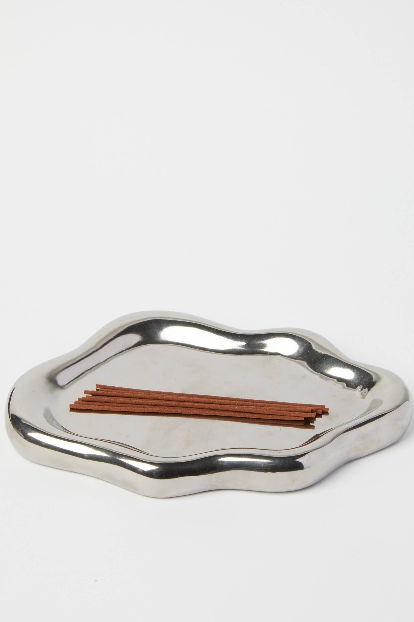 A polished silver metal tray with an organic, wavy scalloped edge, shown on a plain light gray background with a small bundle of brown incense sticks inside.