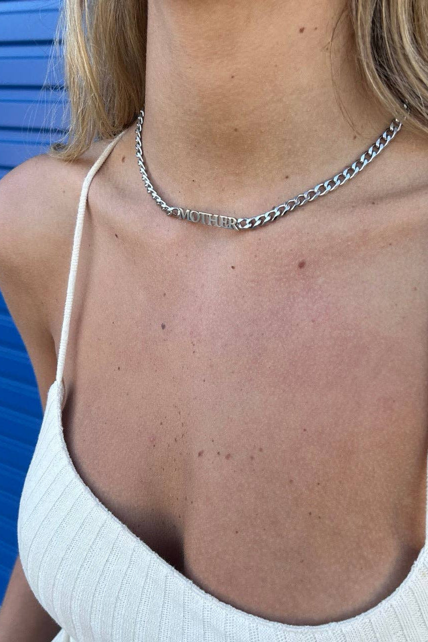 A close-up of a model wearing a silver chain necklace with the word 'mother' spelled out in raised letters, paired with a white ribbed camisole top against a blue corrugated metal background.