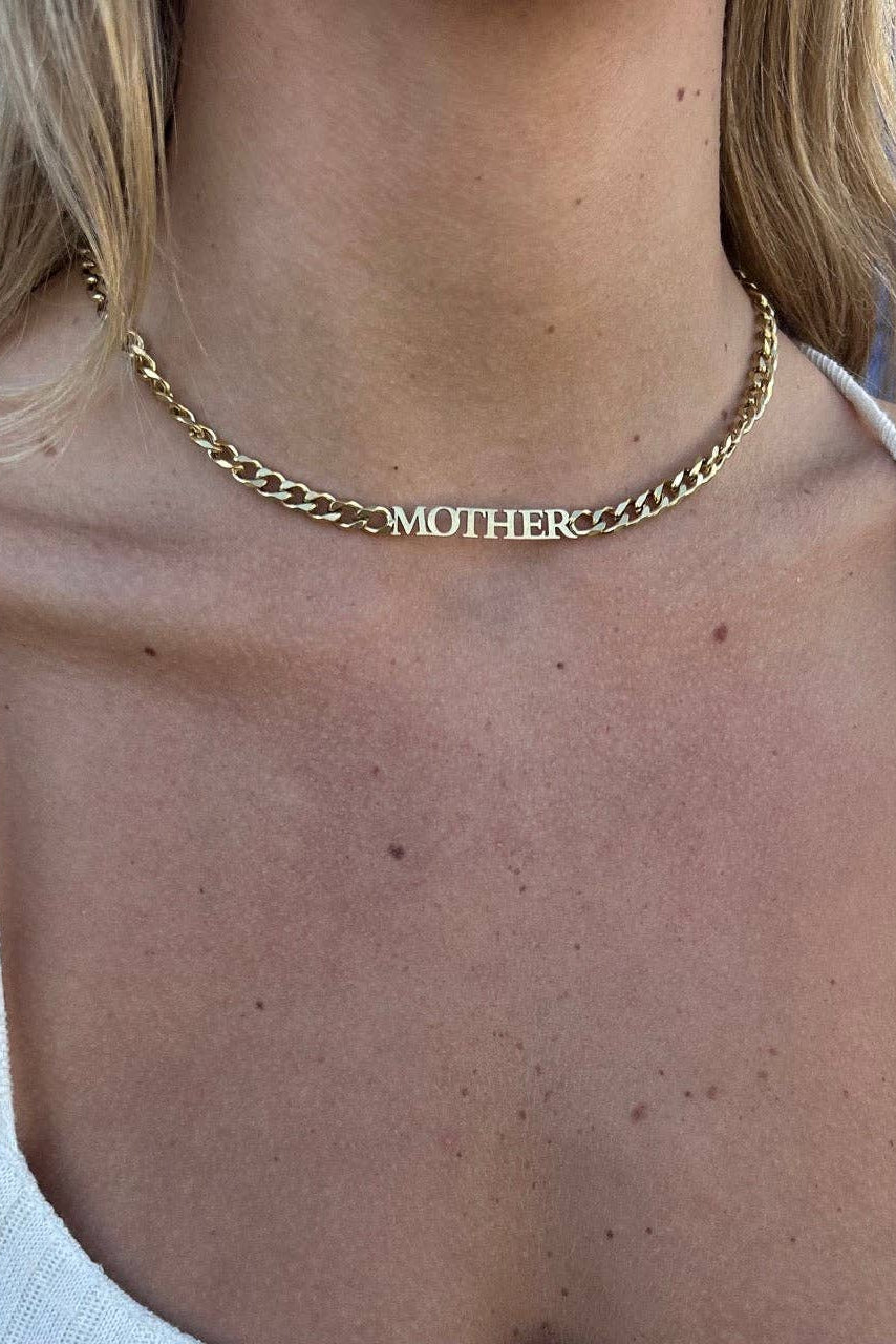 A close-up of a woman wearing a gold chain necklace with the name 'MOTHER' spelled out in block letters across the center.