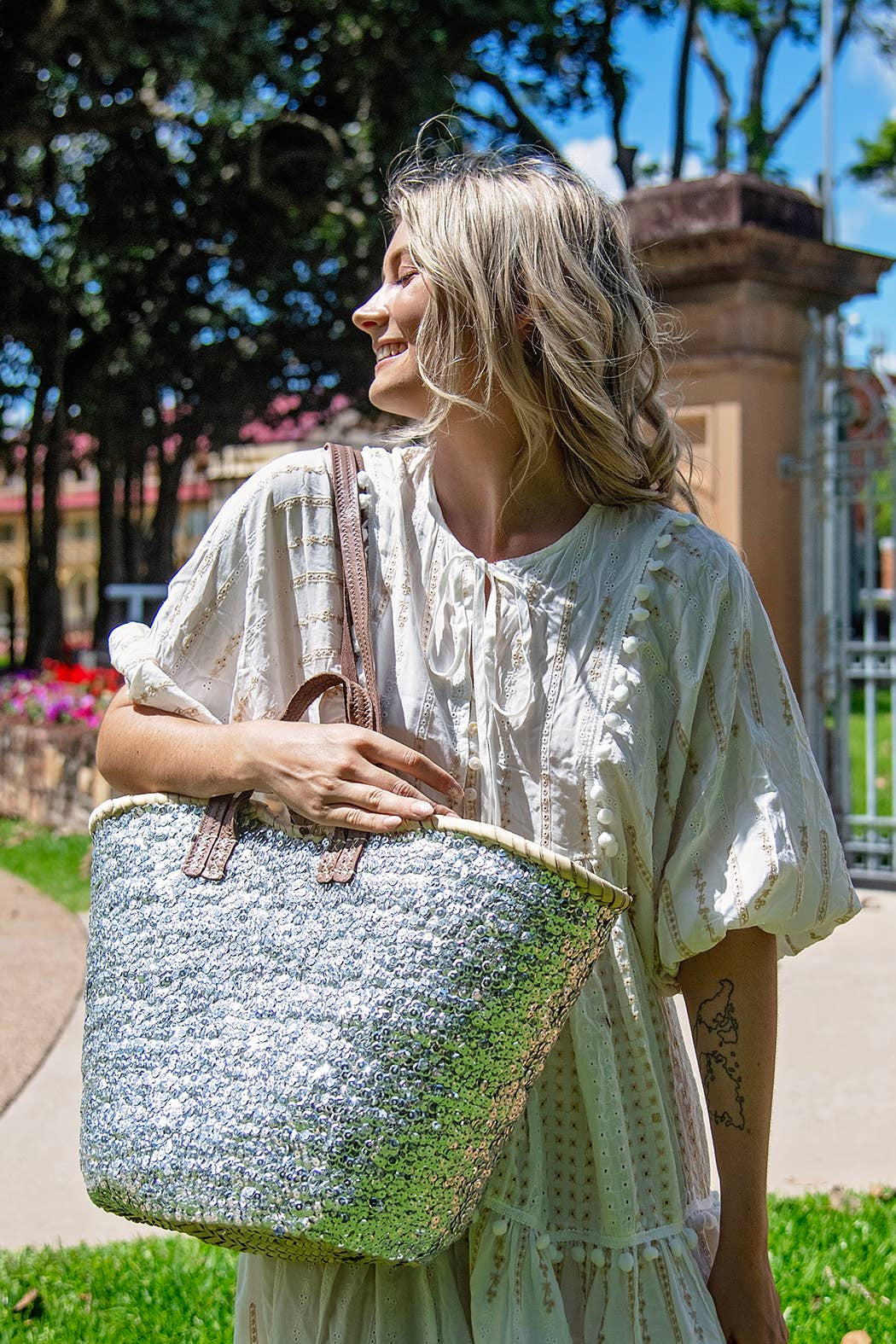 A woman stands outdoors on a sunny day in a grassy park area, holding a large woven straw tote bag with brown leather handles. She is wearing a light-colored, loose-fitting dress.