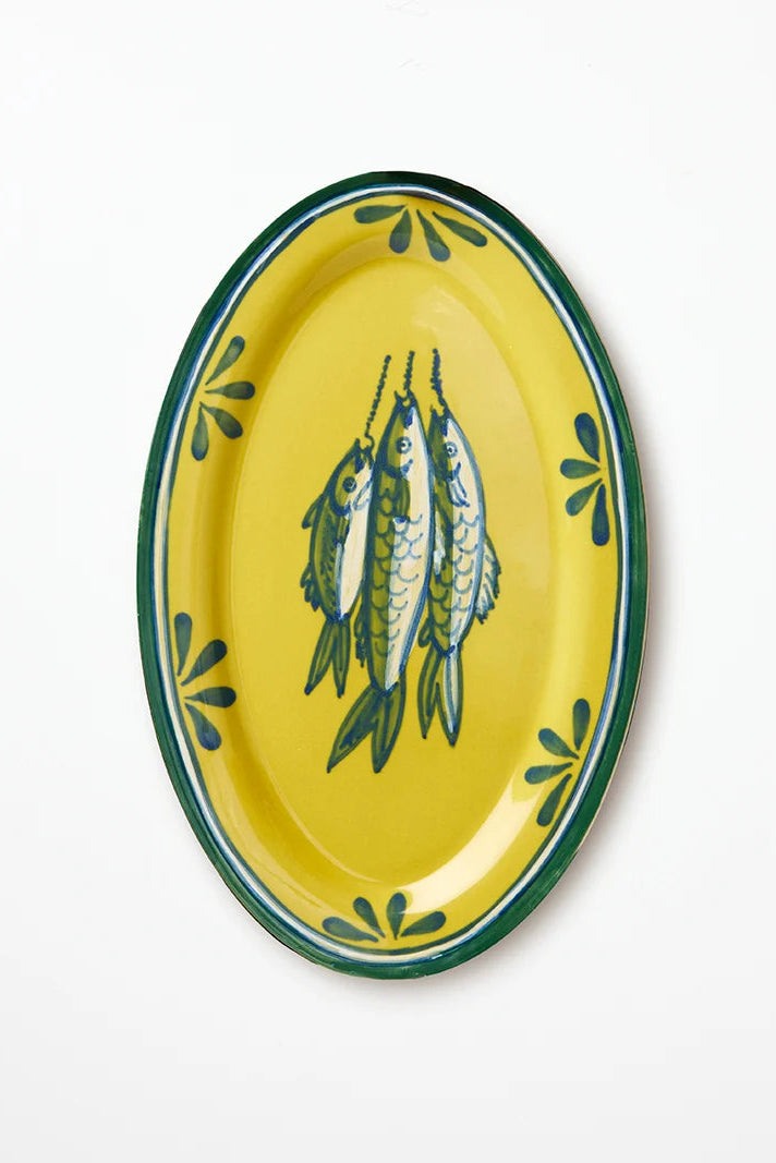 An oval yellow ceramic serving platter featuring a green rim, green leaf-like motifs around the border, and a central illustration of three fish.