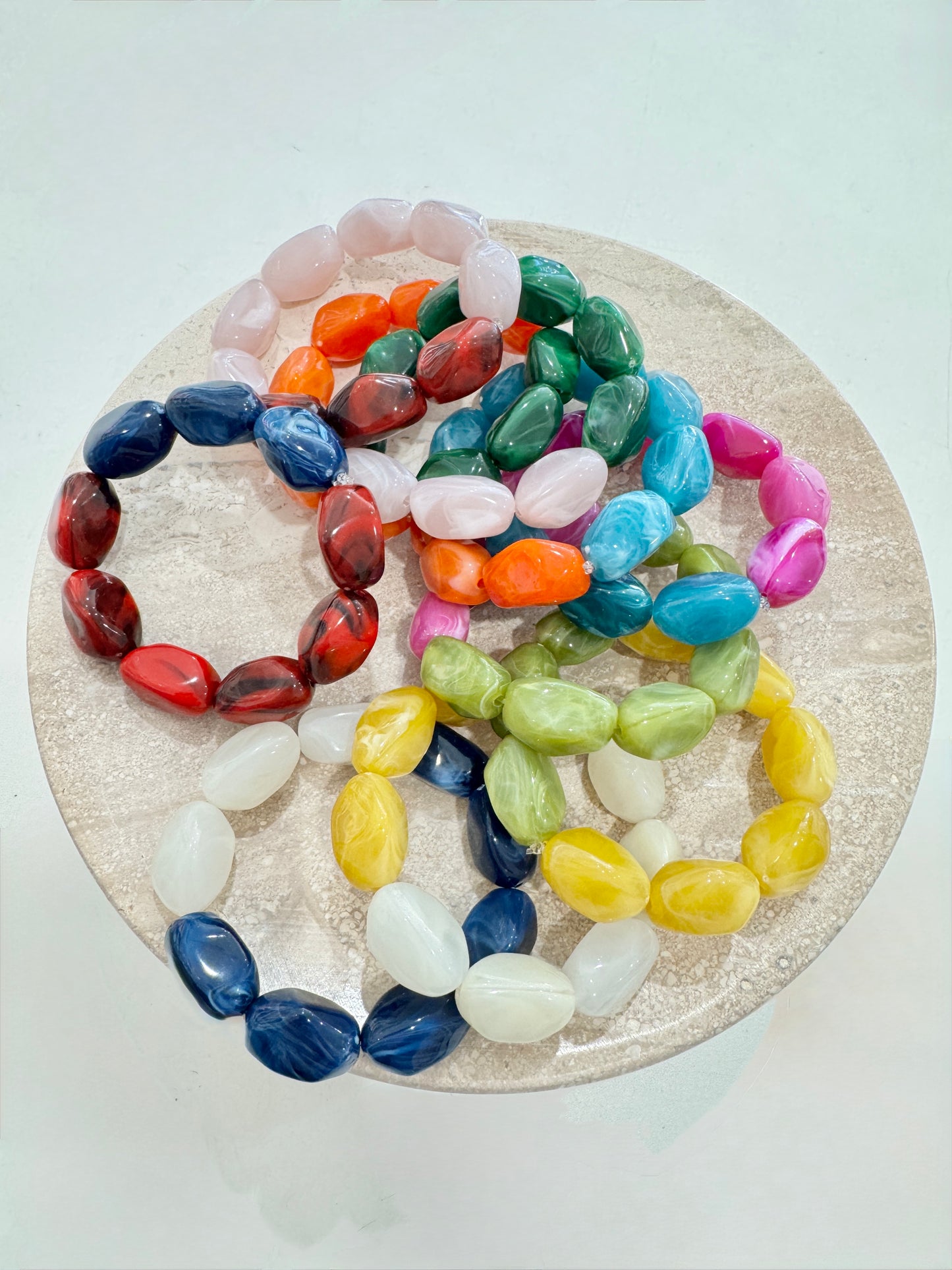 A group of colorful beaded stretch bracelets arranged on a round beige tray against a light background. The bracelets feature smooth, polished oval beads in multiple colors including pink, orange, green, blue, yellow, white, and red.